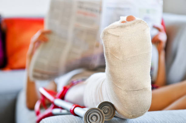 Woman with broken leg lying on sofa and reading newspaper stock photo