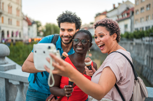 Cheerful friends relishing ice cream, laughter, and city exploration. They capture memories on a bridge, taking selfies with an analog camera.