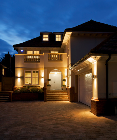 a late evening exterior view of a luxury new home with all internal and outside lights switched on. a beautifully built house with steps leading up to a large white double front door. A larged paved area in the foreground is illuminated by the spotlights on the house.