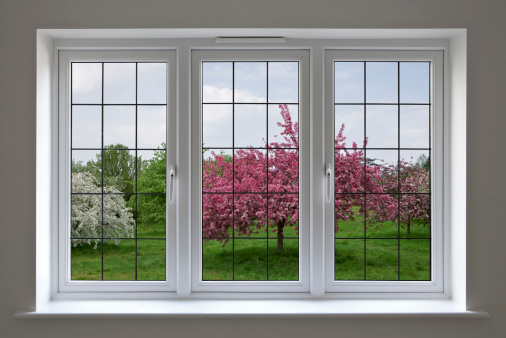Beautifully made white wood framed windows with leaded glass panels, surrounded by a light grey wall. The view through the window is over an apple orchard under a cloudy blue sky. This is a composite of two images.