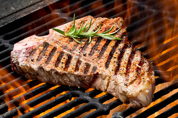 T-Bone Steak on Grill with Flames T-Bone Steak on Grill with Flames on an old fashioned charcoal barbecue.  Steak is garnished with a sprig of rosemary, is seasoned perfectly and the grill marks are perfect. t bone steak stock pictures, royalty-free photos & images