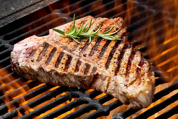 T-Bone Steak on Grill with Flames on an old fashioned charcoal barbecue.  Steak is garnished with a sprig of rosemary, is seasoned perfectly and the grill marks are perfect.