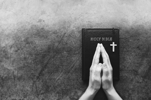 Christian background with the cross of Jesus Christ on an old table and people putting their hands together in meditation, praying and worshiping on the holy Bible.