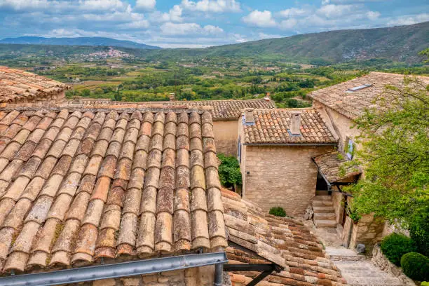 In historic Lacoste, Provence, traditional old stone houses with tiled roofs overlook the lush French countryside, with the village of Bonnieux visible in the distance.