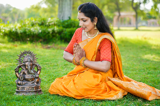 Happy young indian woman wearing saree sitting worshiping lord Ganesha idol outdoor at park celebrating Ganesh Chaturthi festival.