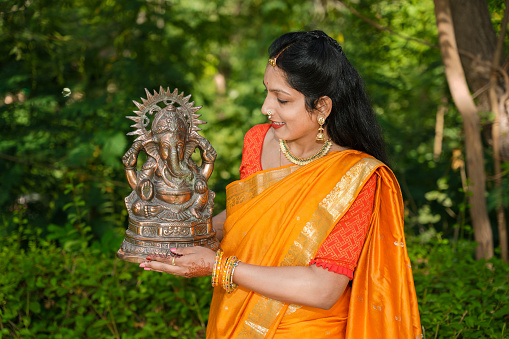 Happy young indian woman wearing saree holding lord Ganesha idol outdoor at park celebrating Ganesh Chaturthi festival.