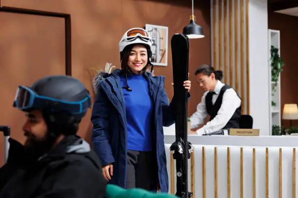 Image showcasing excited female traveler with wintersports equipment at ski resort. Portrait of young asian woman having ski helmet winter jacket and skiing skis is standing in hotel reception.