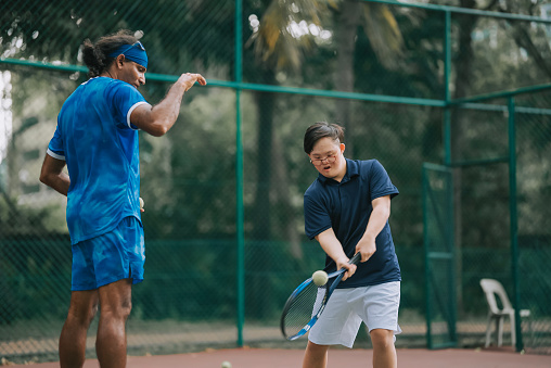 Asian Chinese Down syndrome man learning from male coach trainer playing tennis during weekend morning