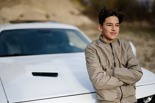 Teenager boy stand near white muscle car. Young teen driver.
