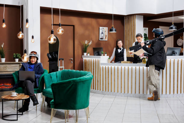 Image shows asian woman with a snowboard seated on sofa using laptop while young man stands at front desk with skiing skis. Visitors in snow clothing and wintersports gear in hotel lobby for check-in.