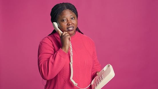 African american woman in stylish outfit having a conversation through handset phone. Female fashion blogger talking on a landline telephone in front of isolated pink background.