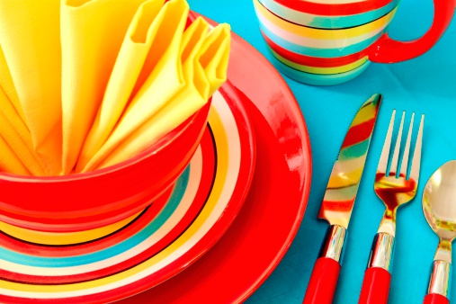 Place setting with bright red, yellow, and turquoise striped dishes and yellow napkin on turquoise tablecloth. Horizontal image.