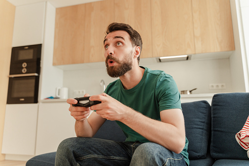Portrait of excited handsome bearded male gamer playing video game, holding joystick, sitting on comfortable sofa in kitchen at home, closeup. Smiling man wearing casual clothes online having fun