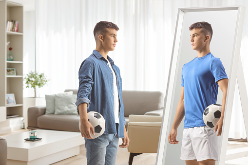 Young man holding a football and looking his self as a football player in a mirror at home