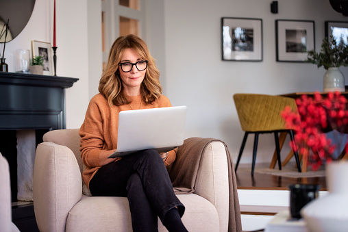 Portrait of  blond haired woman wearing casual clothes while sitting in an armchair at home. Attractive female using laptop for work or having video call. Home office.