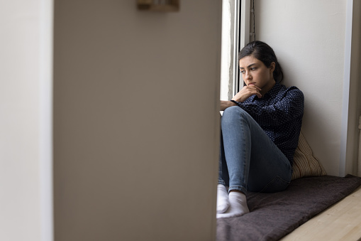 Frustrated pensive Indian woman sits on windowsill and looks out window, suffer from apathy, thinks about relationship problems, experiencing emotional crisis, depression, stress. Break up, abortion