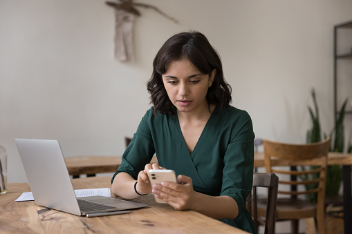 Serious millennial female holding smartphone seated at desk in workplace, distracted from laptop freelance work, learn new mobile business application, read received notice, use e-bank check incomes