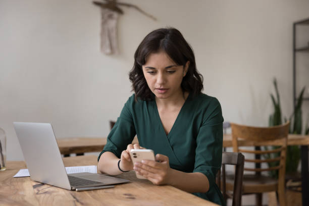 Serious millennial female holds smartphone seated at desk stock photo