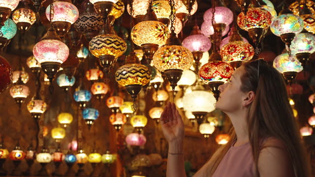 A young woman is examining antique colorful Turkish lamps hanging from the ceiling in a shop.