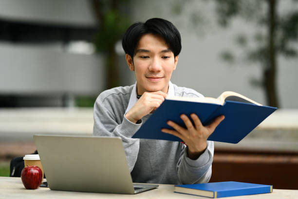 istock Smiling man reading book or studying for exam, doing homework remotely at a table in front of university building 1660931180