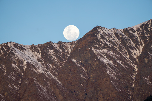 Luminous moon dominates the sky, casting light on a rugged mountain below. This celestial scene captures the serenity of full moonrise over the dramatic landscape. Queenstown, New Zealand