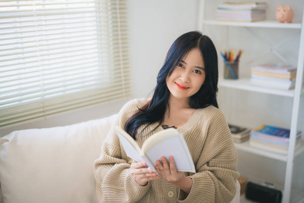 Joyful happy asian woman smiling and reading book while sitting on couch sofa in living room at home. Asian women relax reading book smile at sofa In the house. Activity hobby at house concept. alegre mujer asiática feliz sonriendo y leyendo un libro mientras está sentada en el sofá del sofá en la sala de estar de su casa. las mujeres asiáticas se relajan leyendo el libro sonríen en el sofá de la casa. concepto de actividad hobby en casa.