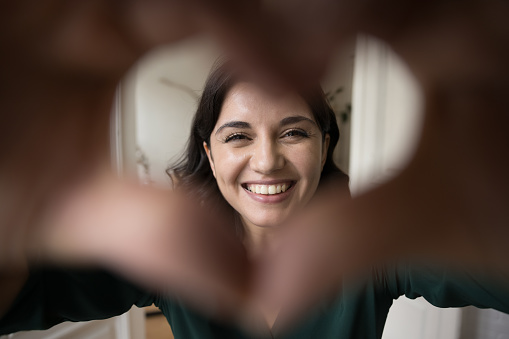 Close up face Armenian overjoyed woman show symbol of love with joined fingers, sign of happiness and romance, look through heart shaped frame, charity, volunteering, professional medical services ad