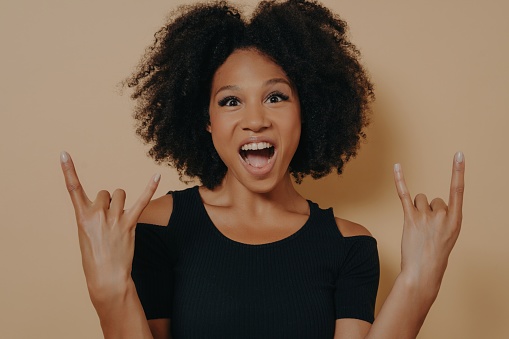 Energetic African American Woman in Black T-shirt Shouts with Wild Expression, Rock-n-Roll Hand Gesture, Isolated on Beige Studio Background, Offering Copy Space
