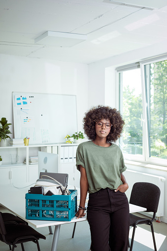 Confident afro american young woman wearing green t-shirt standing in the office next to e-waste recycling bin, looking at camera.