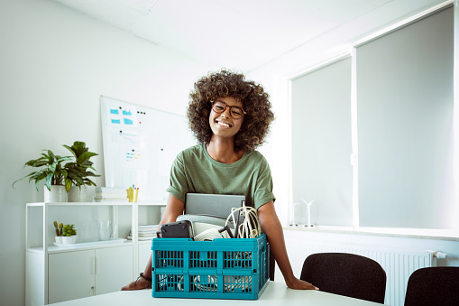 Afro american young woman wearing green t-shirt standing in the office next to e-waste recycling bin and smiling at camera.