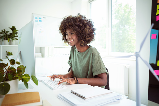 Afro american young woman wearing green t-shirt sitting at the desk in the office with defocused wind turbine model in the foreground, working on computer.