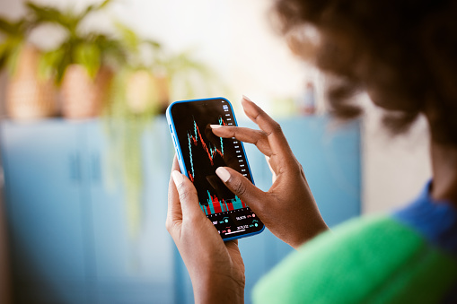 Afro american woman using smart phone at home, watching stock charts. Close up of hands, unrecognizable person.
