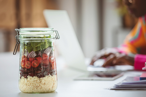 Focus on homemade jar salad with defocused afro american young woman working on laptop in the office.