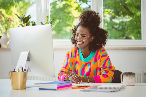 Afro american young woman wearing colorful sweater sitting at the desk at home and using computer, having video call. Female freelancer working at home.