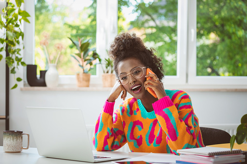 Afro american young woman wearing colorful sweater sitting at the desk at home, using laptop and talking on mobile phone. Female freelancer working at home.