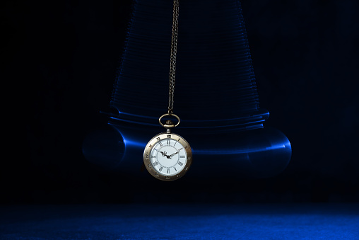 Hypnosis session. Vintage pocket watch with chain swinging over surface on dark background among faded clock faces, magic motion effect