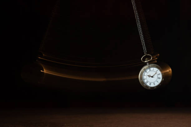 istock Hypnosis session. Vintage pocket watch with chain swinging over surface on dark background among faded clock faces, magic motion effect 1661031376