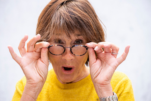 Shocked mature caucasian woman with open mouth removing glasses and checking something astonishing
