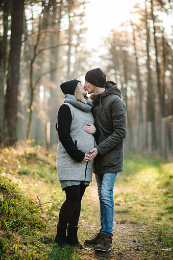 Pregnant woman kissing man stands and hug in background golden leaves in forest at sunset. Young female hug male spending time together in nature. Happy couple walk in autumn park. Autumn family photo