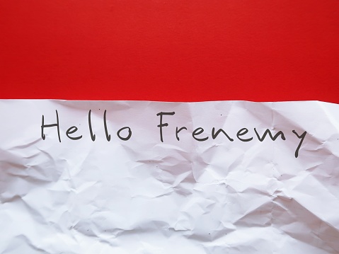 Crumpled paper on red background with handwriien text - Hello Frenemy - person who combines the characteristics of friend and enemy, rival pretends to be a friend