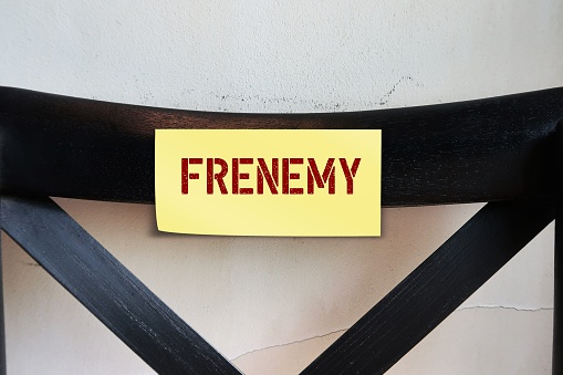 Chair back with yellow stick note with text - FRENEMY - person who combines the characteristics of friend and enemy, rival pretends to be a friend