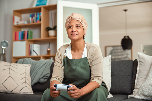 In a cozy and familiar setting, a mature woman is engrossed in playing video games while seated on her sofa at home. Her focus demonstrate that age is no barrier to enjoying the world of gaming.