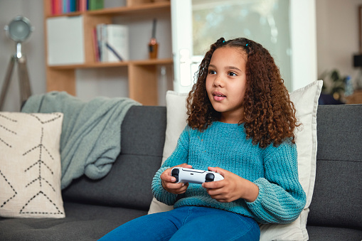 A young girl is engrossed in playing video games on the sofa at home. Her eyes are fixed on the screen, and she's eagerly manipulating the controller to navigate through the virtual world. The room is filled with the soft glow of the screen as she enjoys her gaming session.