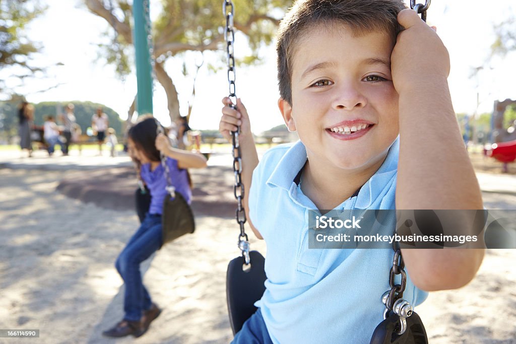 Grinning boy on a swing at a park with a girl in the back Boy And Girl Playing On Swing In Park Smiling To Camera Child Stock Photo