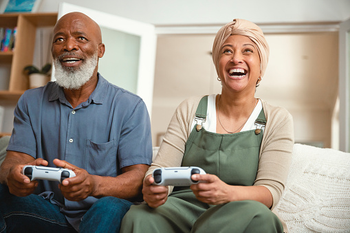 In the comfort of their home, a senior couple is seated together on their sofa, sharing a delightful moment playing video games. Their smiles and laughter highlight the joy and togetherness they find in this shared activity, showcasing that love knows no age when it comes to having fun.