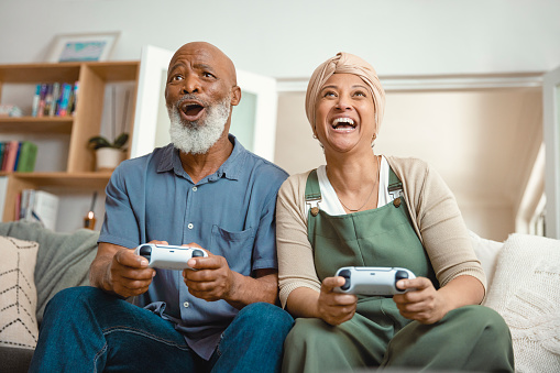 In the comfort of their home, a senior couple is seated together on their sofa, sharing a delightful moment playing video games. Their smiles and laughter highlight the joy and togetherness they find in this shared activity, showcasing that love knows no age when it comes to having fun.