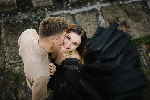 Newlyweds kissing top view. Female embrace male wall in street. Luxury couple. Woman in black long dress hugs man on stairs near ancient palace at sunset. Bride hug groom