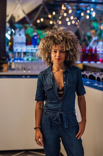 A portrait of a mid adult woman wearing a denim jumpsuit standing in a nightclub in Newcastle, North East England. She is looking at the camera and smiling with her hand slightly in her pocket.