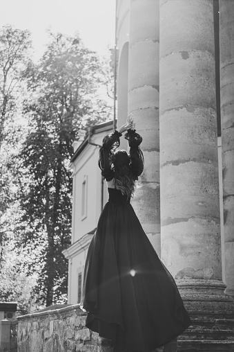 Girl in dress stand on stairs near large columns of ancient temple at sunset. Woman in nature autumn day. Black and white photo. Female walk near Baroque Catholic church in Pidhirtsi. Back view