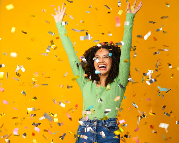 Foto de estúdio de mulher animada comemorando grande vitória banhada em confetes de tinsel no fundo amarelo - foto de acervo